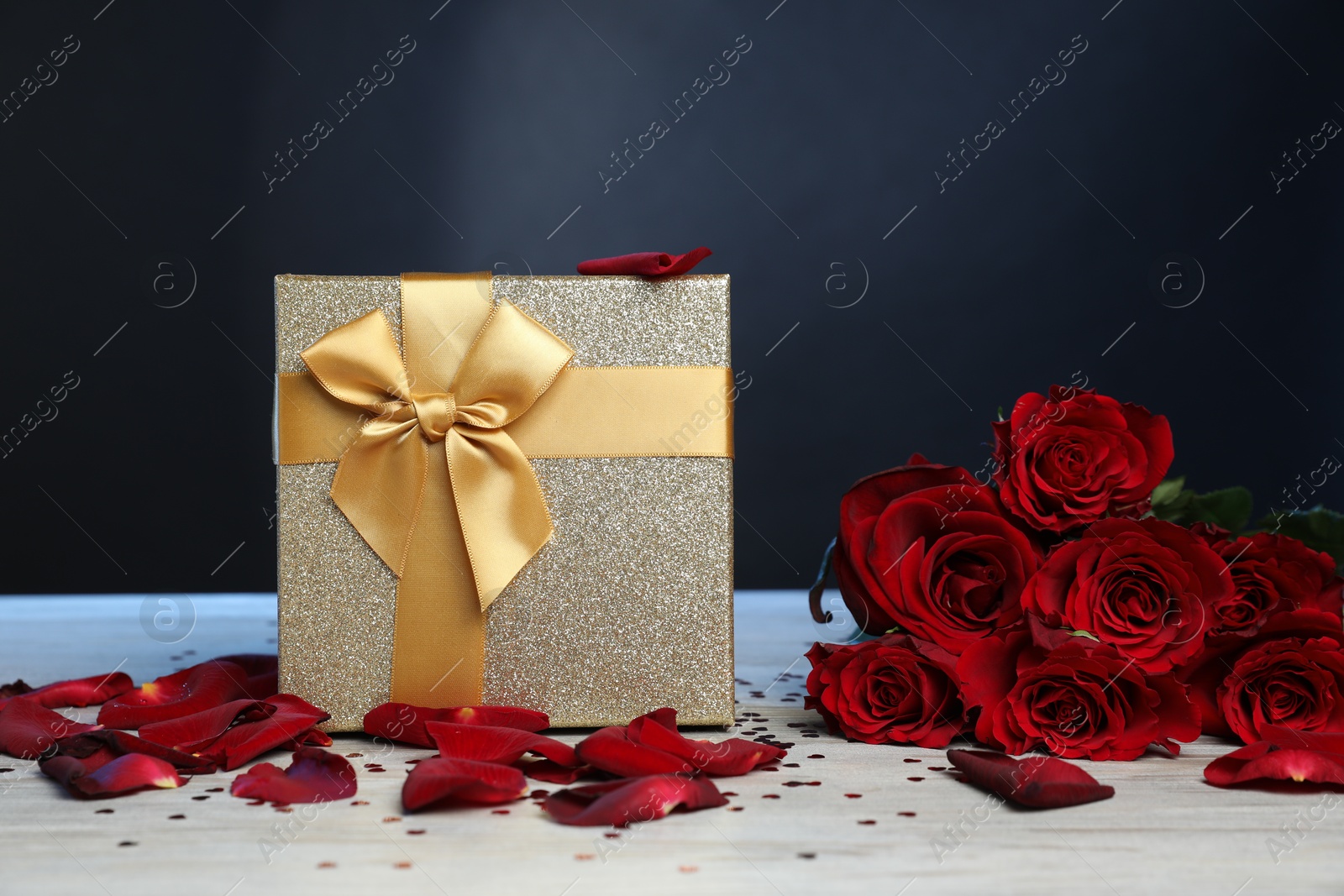 Photo of Beautiful gift box, rose flowers, petals and confetti on white wooden table against dark background, closeup. Romantic present