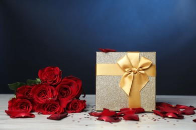 Photo of Beautiful gift box, rose flowers, petals and confetti on white wooden table against blue background, closeup. Romantic present