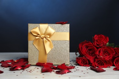 Beautiful gift box, rose flowers, petals and confetti on white wooden table against blue background, closeup. Romantic present