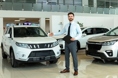 Photo of Happy salesman with clipboard near new cars in salon