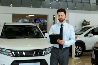 Photo of Happy salesman with clipboard near new cars in salon