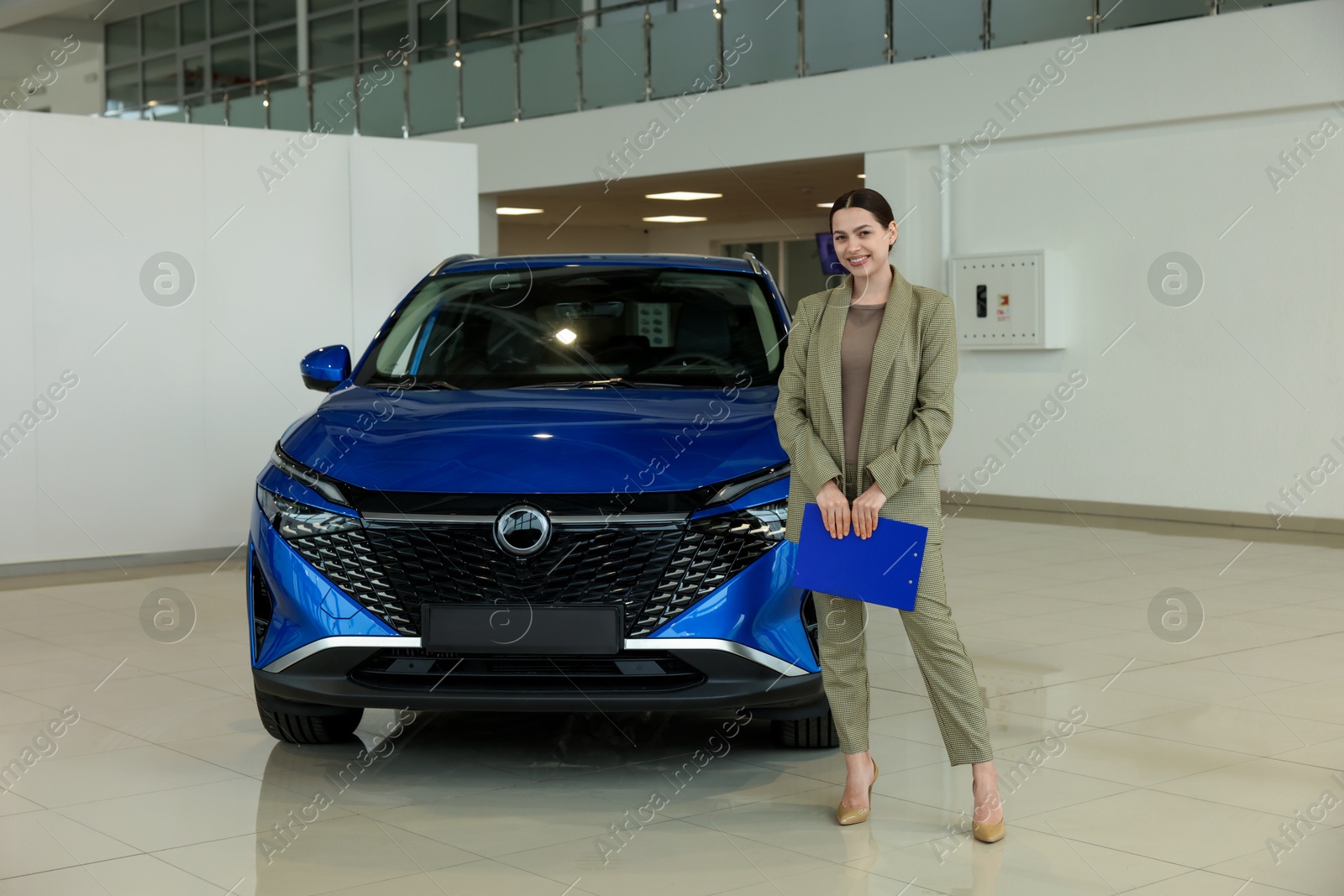 Photo of Happy saleswoman with clipboard near new blue car in salon