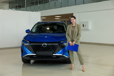 Happy saleswoman with clipboard near new blue car in salon