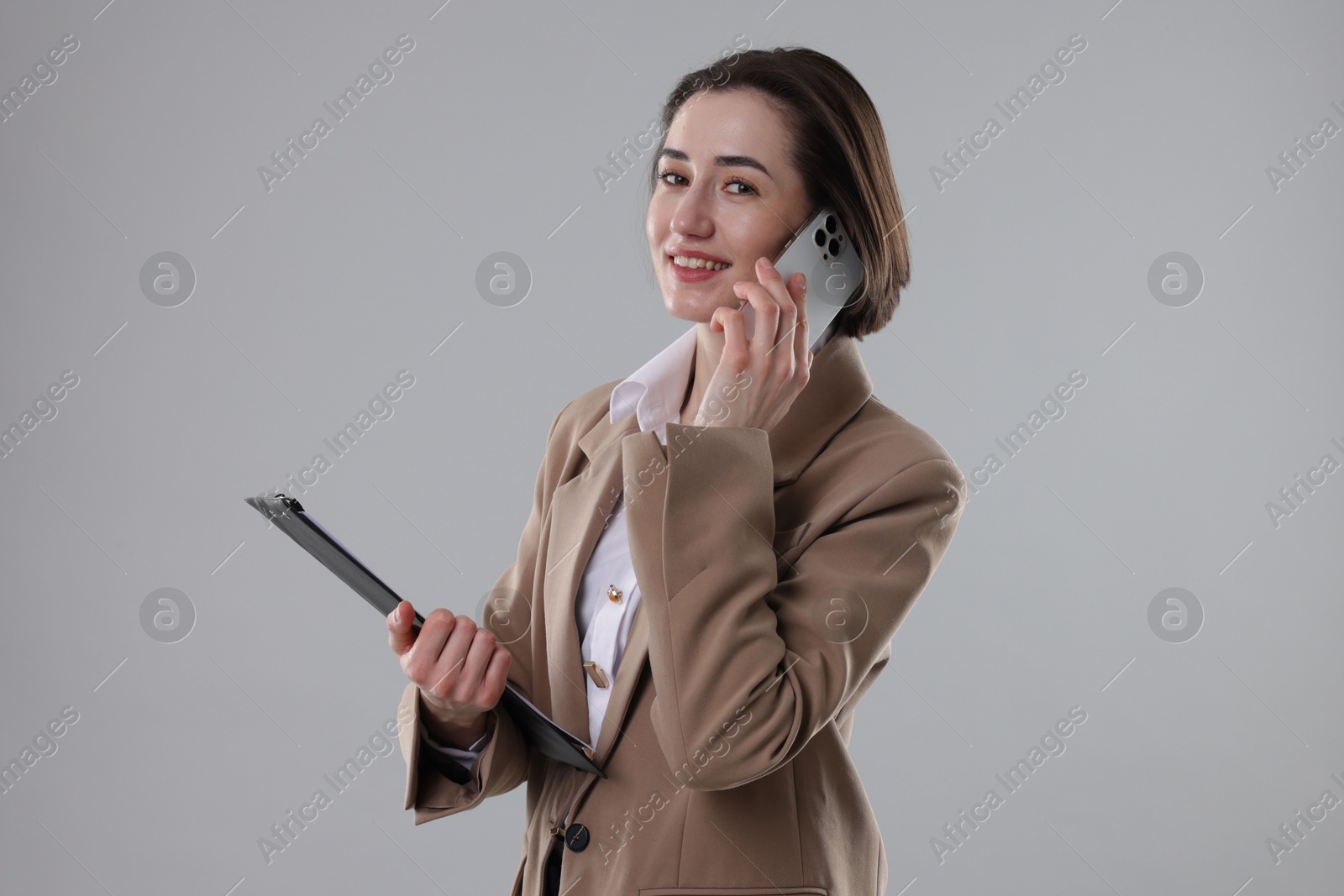 Photo of Smiling secretary with clipboard talking on smartphone against grey background