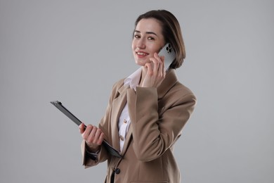 Smiling secretary with clipboard talking on smartphone against grey background