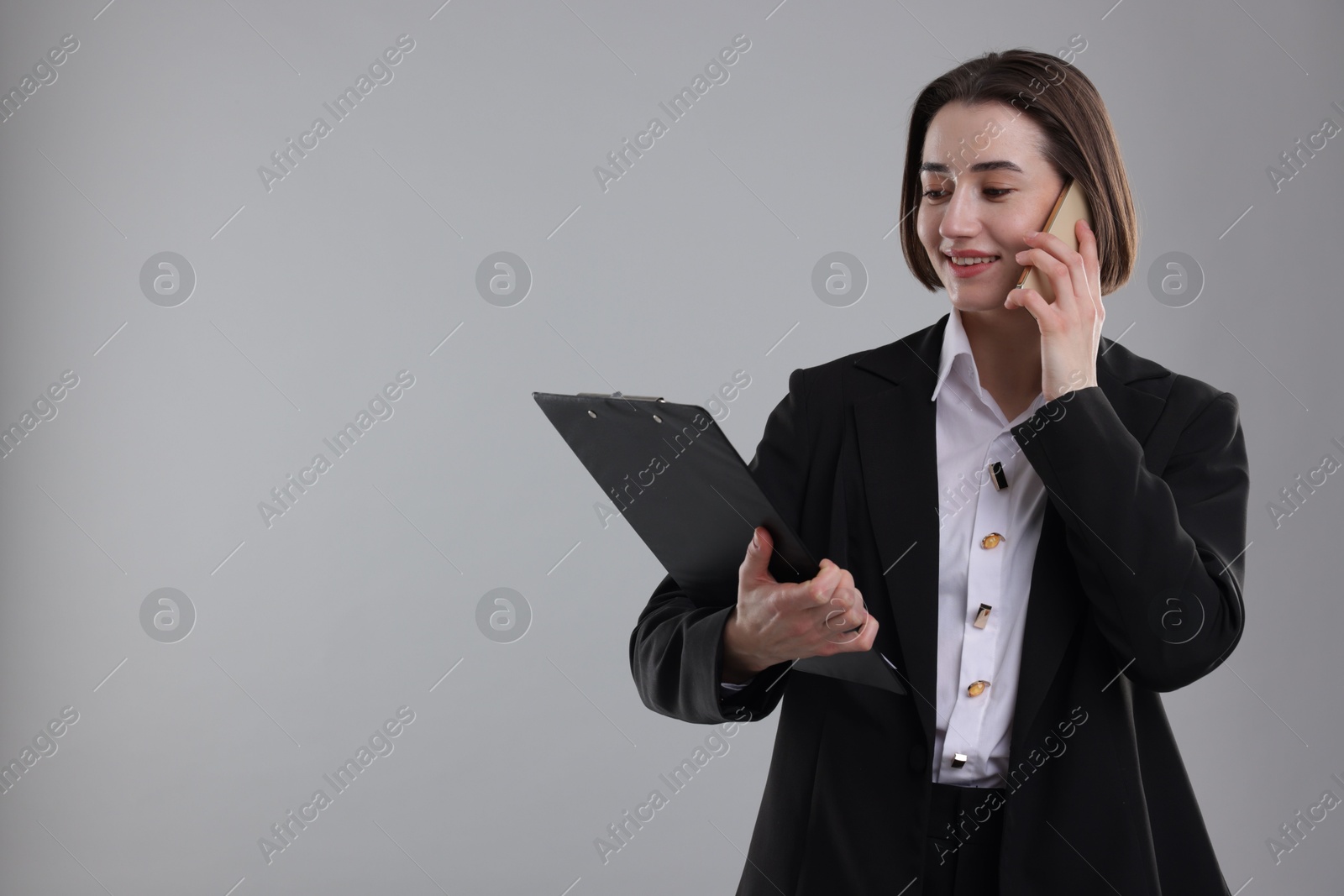 Photo of Smiling secretary with clipboard talking on smartphone against grey background. Space for text