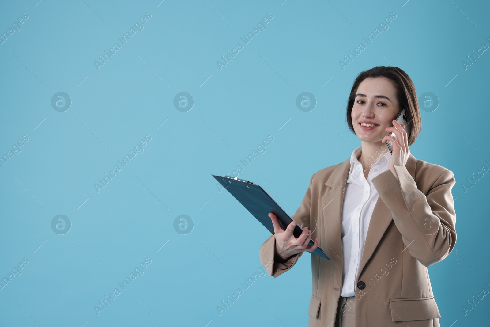 Photo of Smiling secretary with clipboard talking on smartphone against light blue background. Space for text