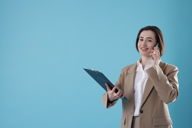 Smiling secretary with clipboard talking on smartphone against light blue background. Space for text