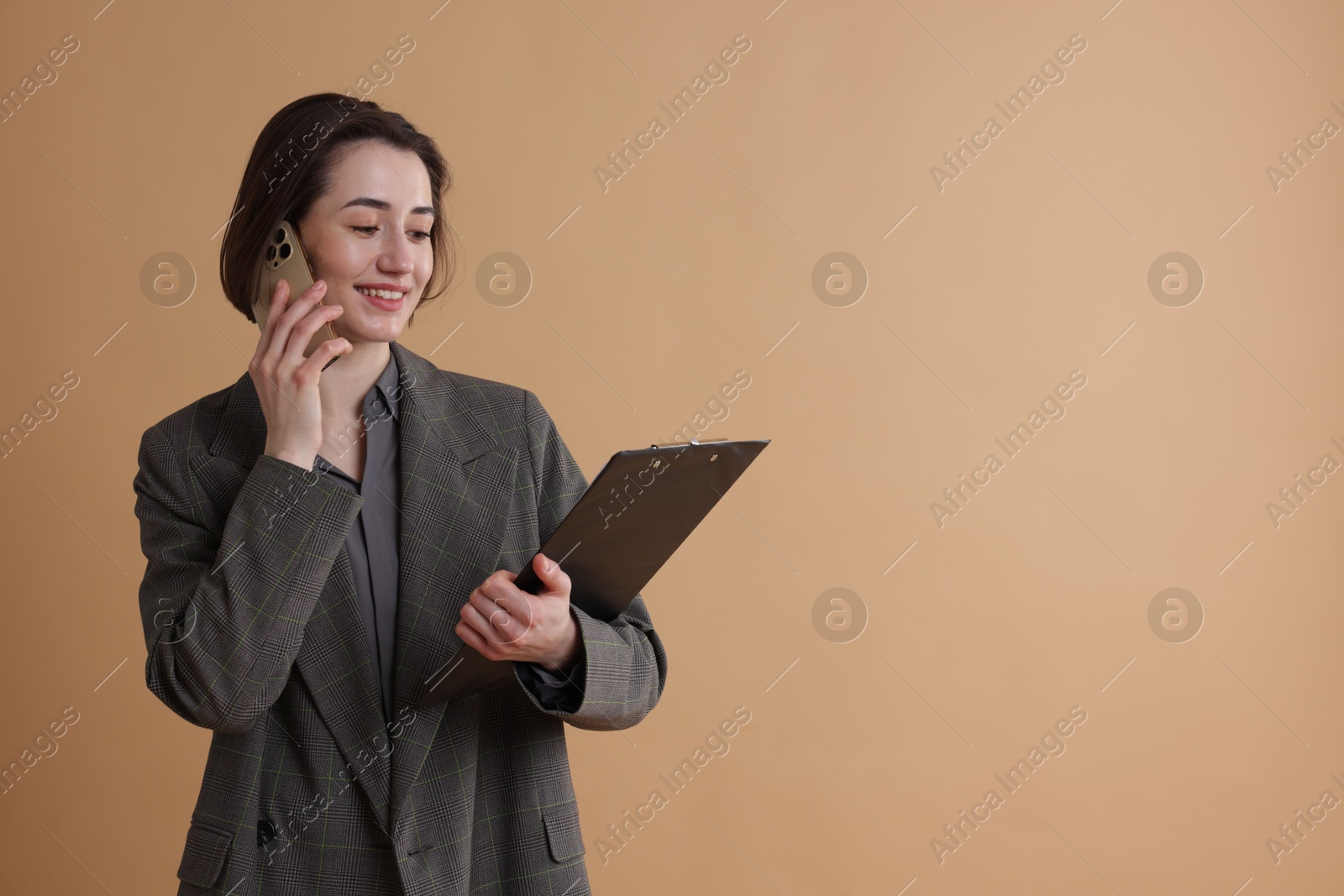 Photo of Smiling secretary with clipboard talking on smartphone against dark beige background. Space for text