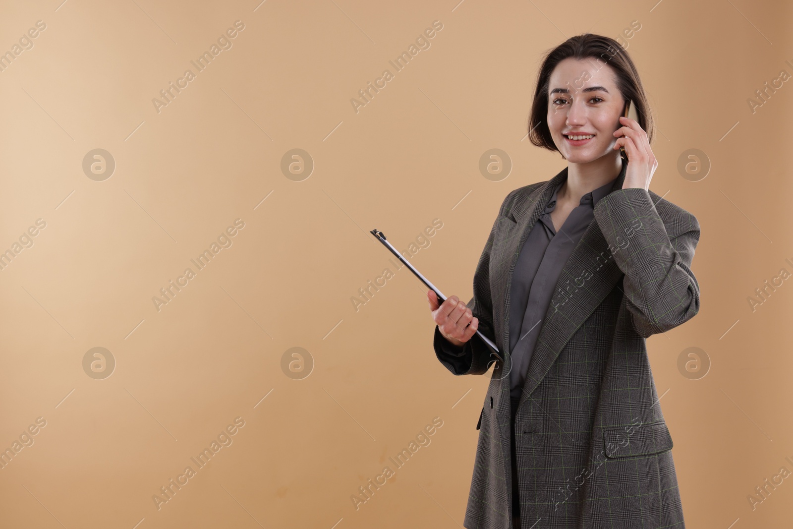 Photo of Smiling secretary with clipboard talking on smartphone against dark beige background. Space for text