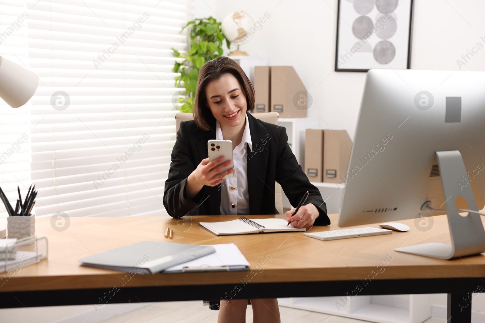 Photo of Smiling secretary with smartphone taking notes at table in office