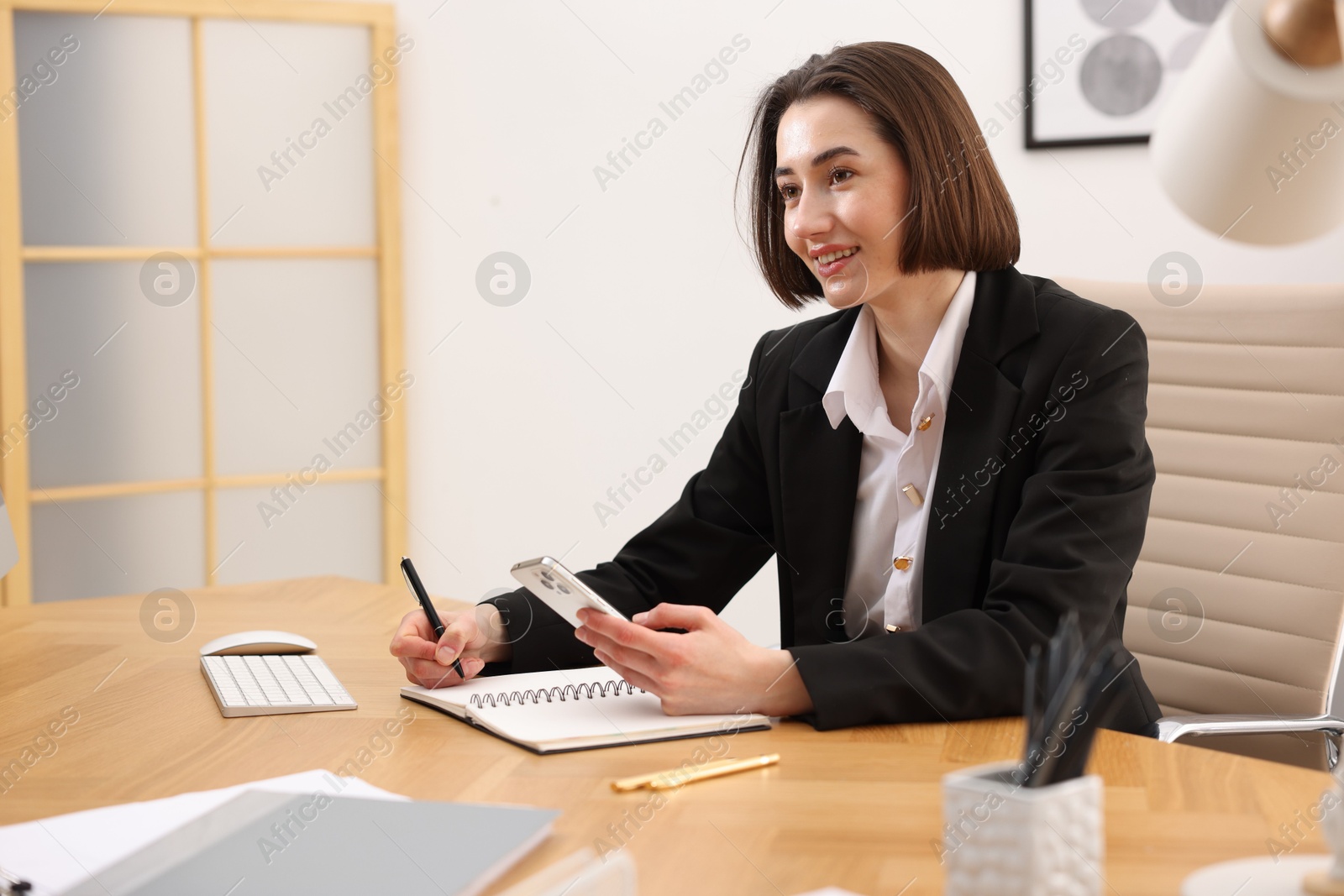 Photo of Smiling secretary with smartphone taking notes at table in office