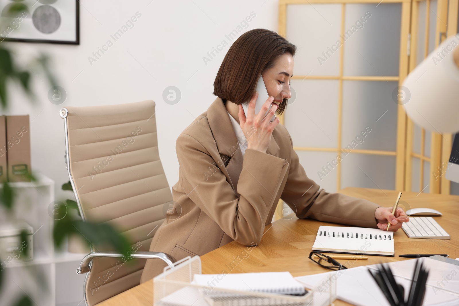 Photo of Smiling secretary talking on smartphone and taking notes at table in office