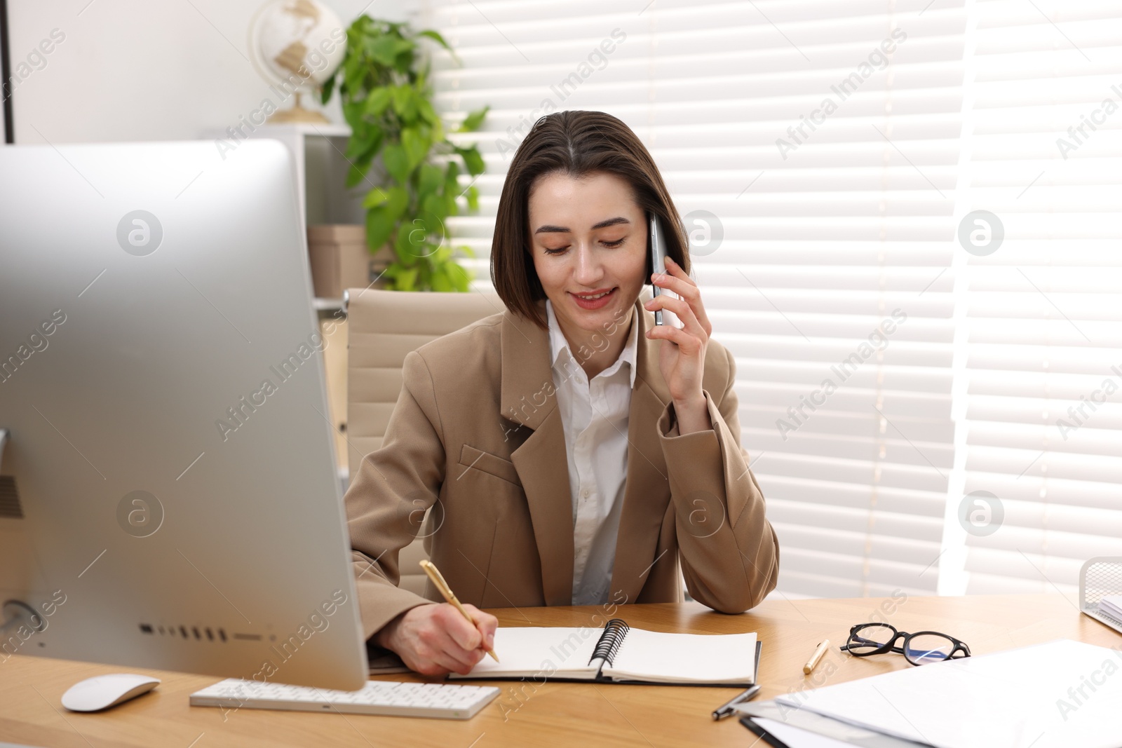 Photo of Smiling secretary talking on smartphone and taking notes at table in office