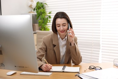 Smiling secretary talking on smartphone and taking notes at table in office