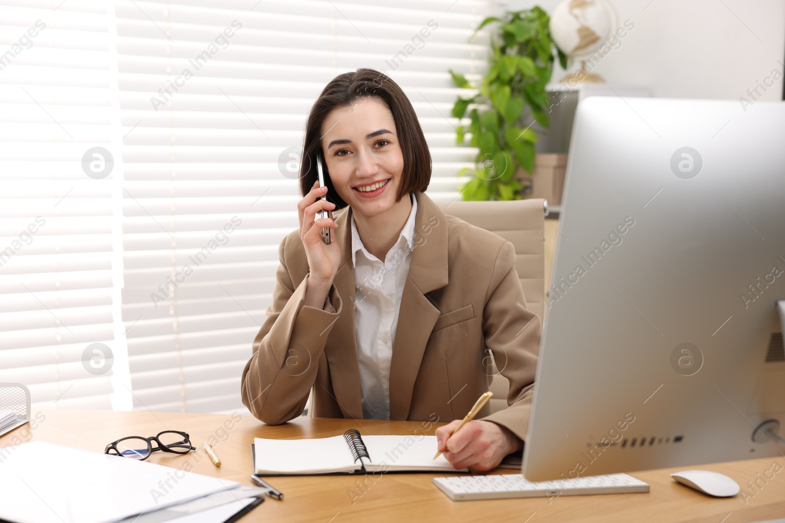 Photo of Smiling secretary talking on smartphone and taking notes at table in office