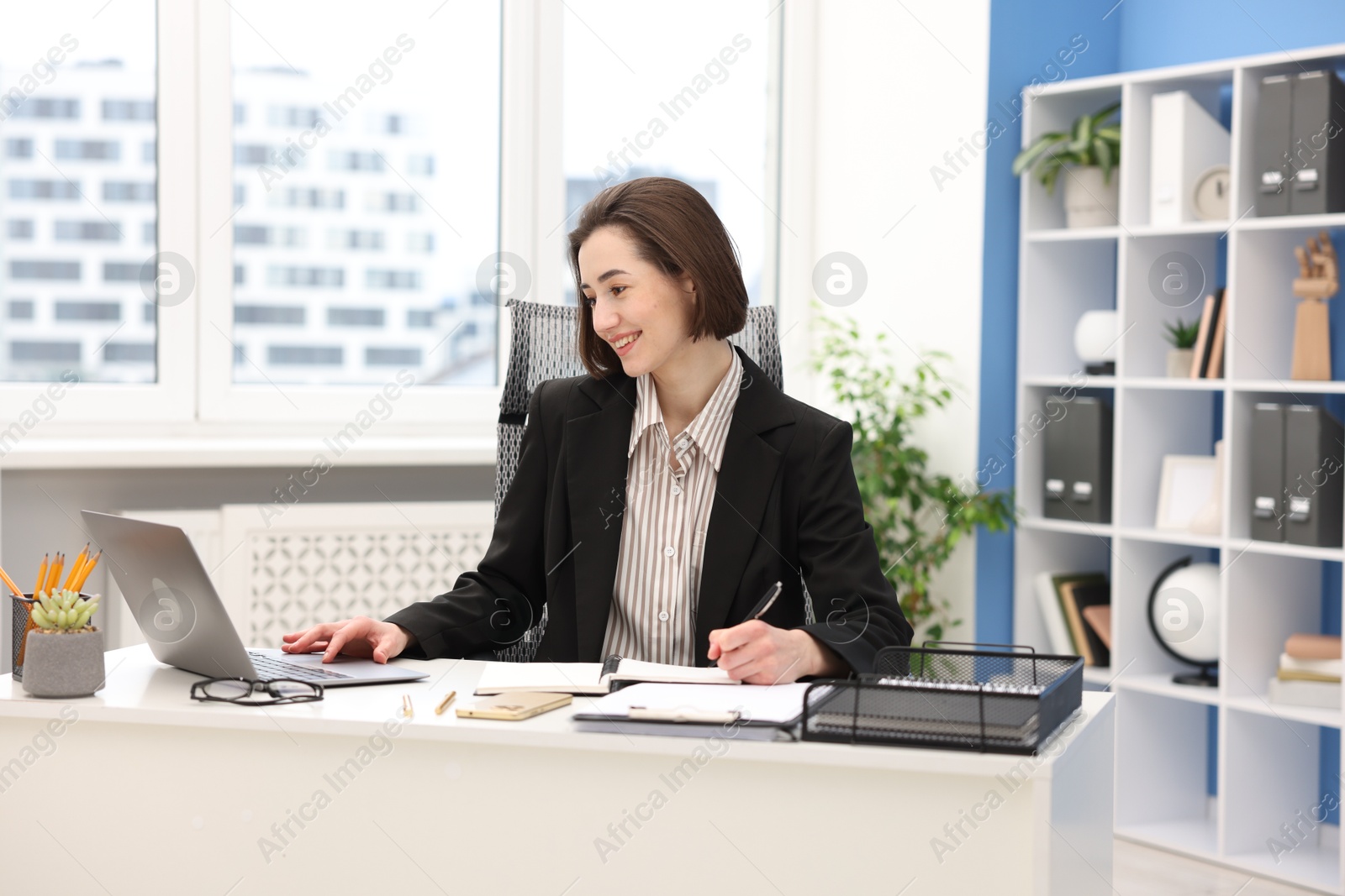 Photo of Smiling secretary taking notes while working on laptop at table in office