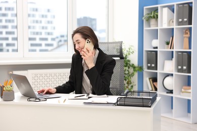 Photo of Smiling secretary talking on smartphone while working with laptop at table in office