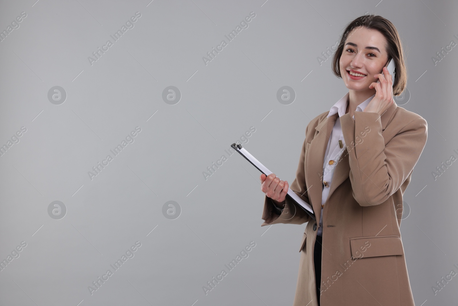 Photo of Smiling secretary with clipboard talking on smartphone against grey background. Space for text