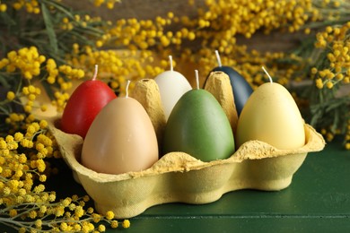 Photo of Colorful egg-shaped candles in carton and mimosa flowers on green wooden table, closeup. Easter decor