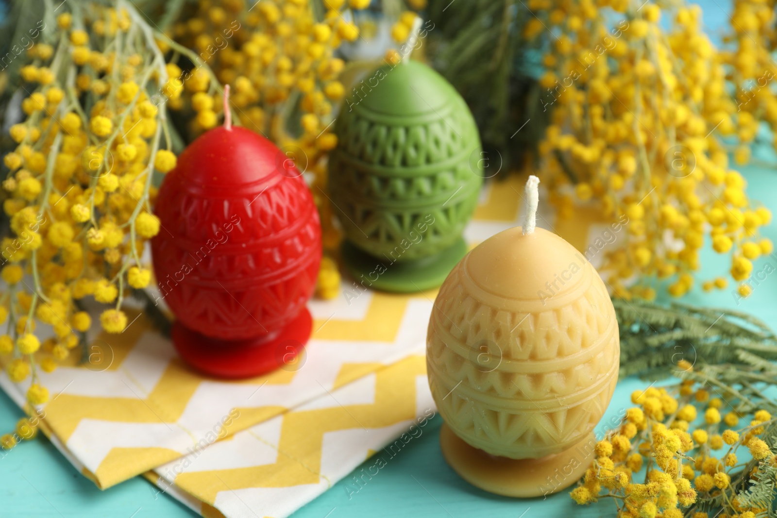 Photo of Colorful egg-shaped candles and mimosa flowers on turquoise table, closeup. Easter decor