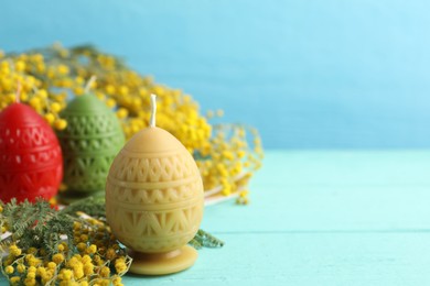 Photo of Colorful egg-shaped candles and mimosa flowers on turquoise wooden table against light blue background, closeup with space for text. Easter decor