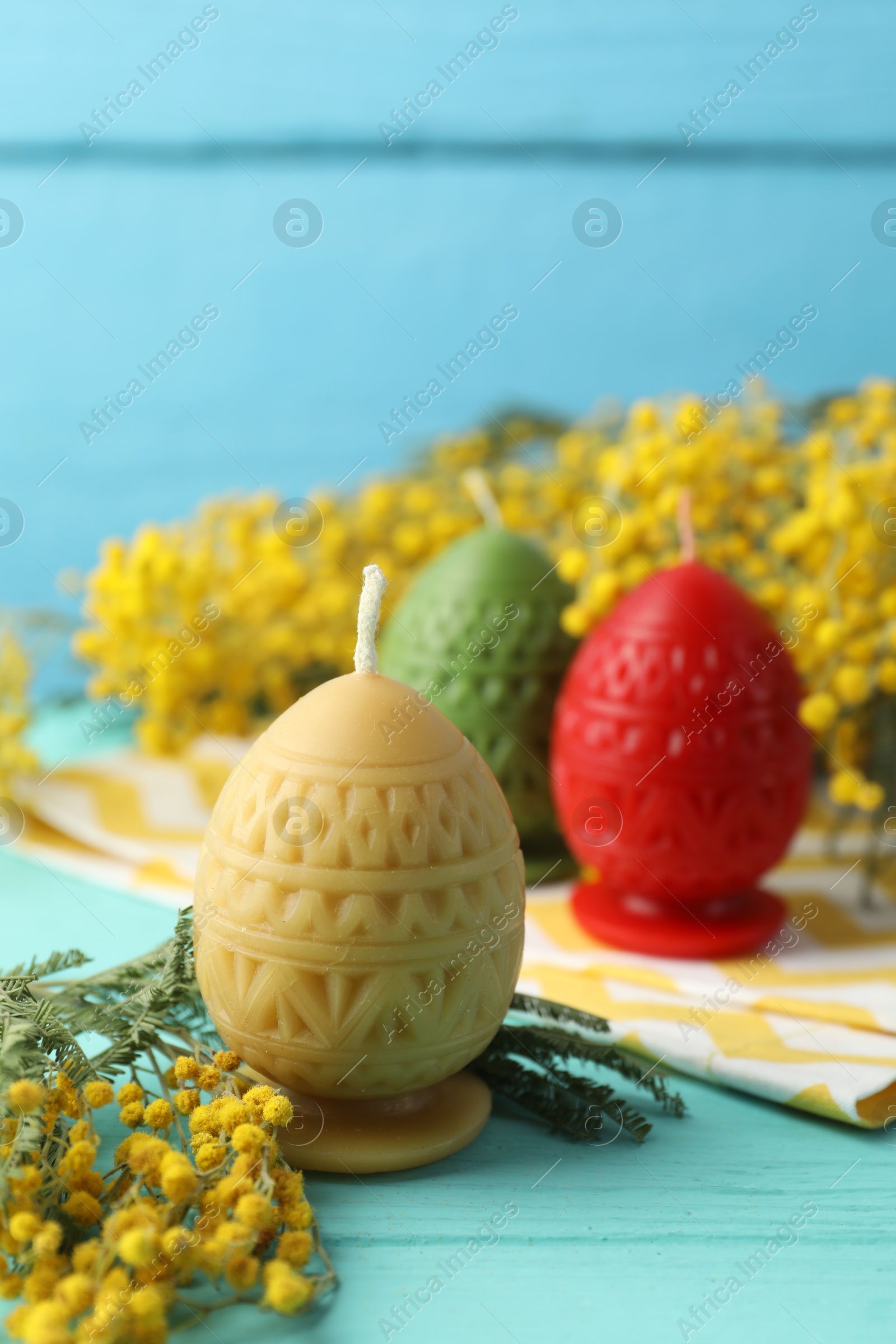 Photo of Colorful egg-shaped candles and mimosa flowers on turquoise wooden table against light blue background, closeup. Easter decor