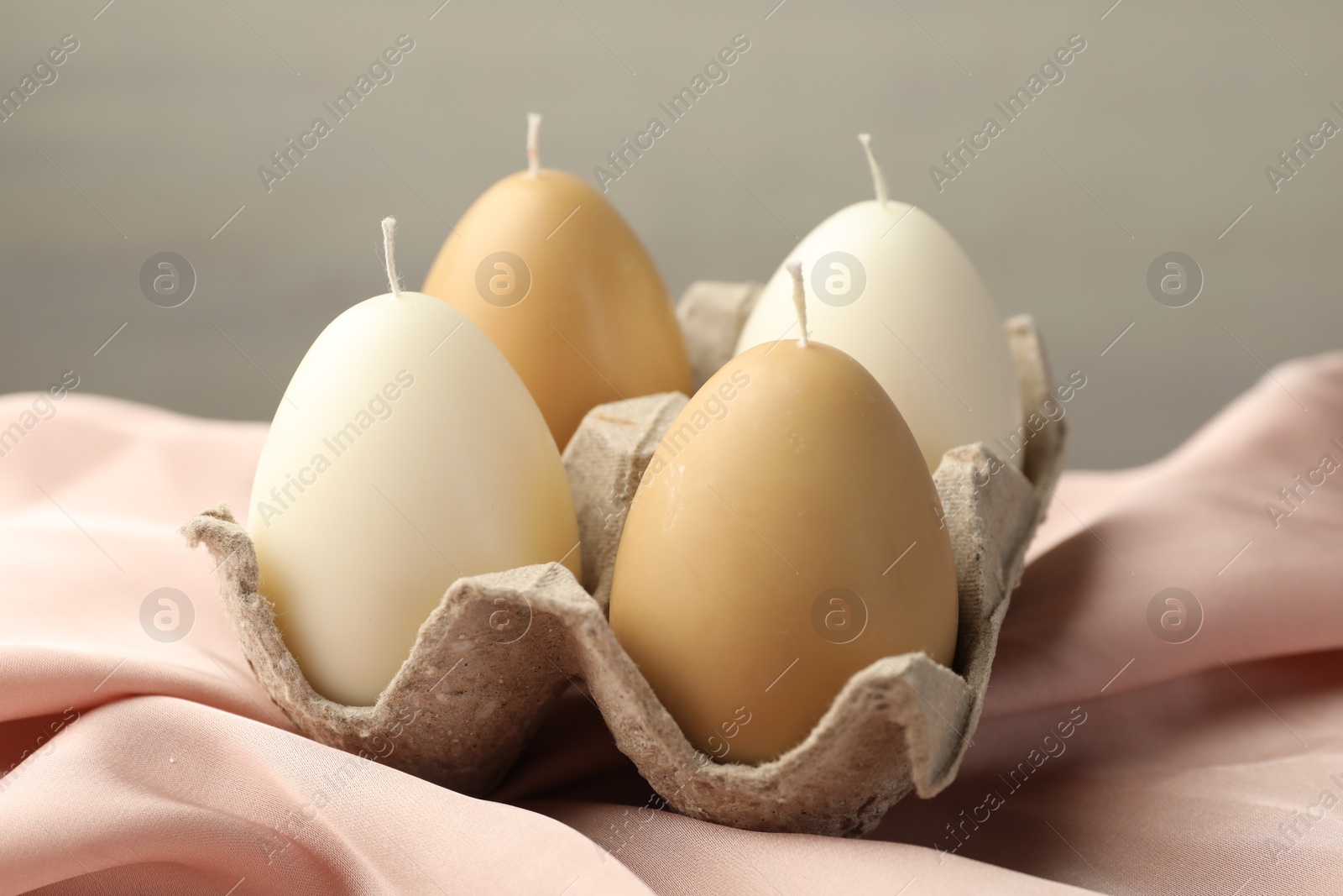 Photo of Egg-shaped candles in carton beige fabric against light gray background, closeup. Easter decor