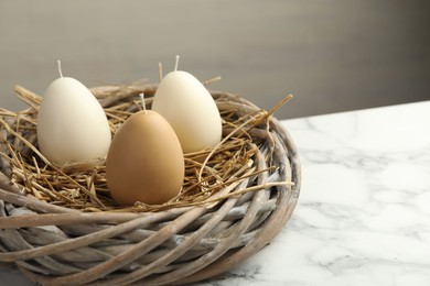 Photo of Egg-shaped candles in nest on white marble table, closeup. Easter decor