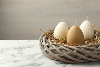 Photo of Egg-shaped candles in nest on white marble table against light gray background, space for text. Easter decor