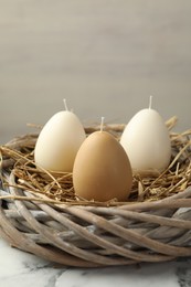 Photo of Egg-shaped candles in nest on white marble table against light gray background, closeup. Easter decor