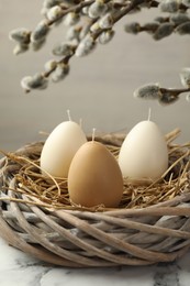 Photo of Egg-shaped candles in nest on white marble table and willow branches against light background, closeup. Easter decor