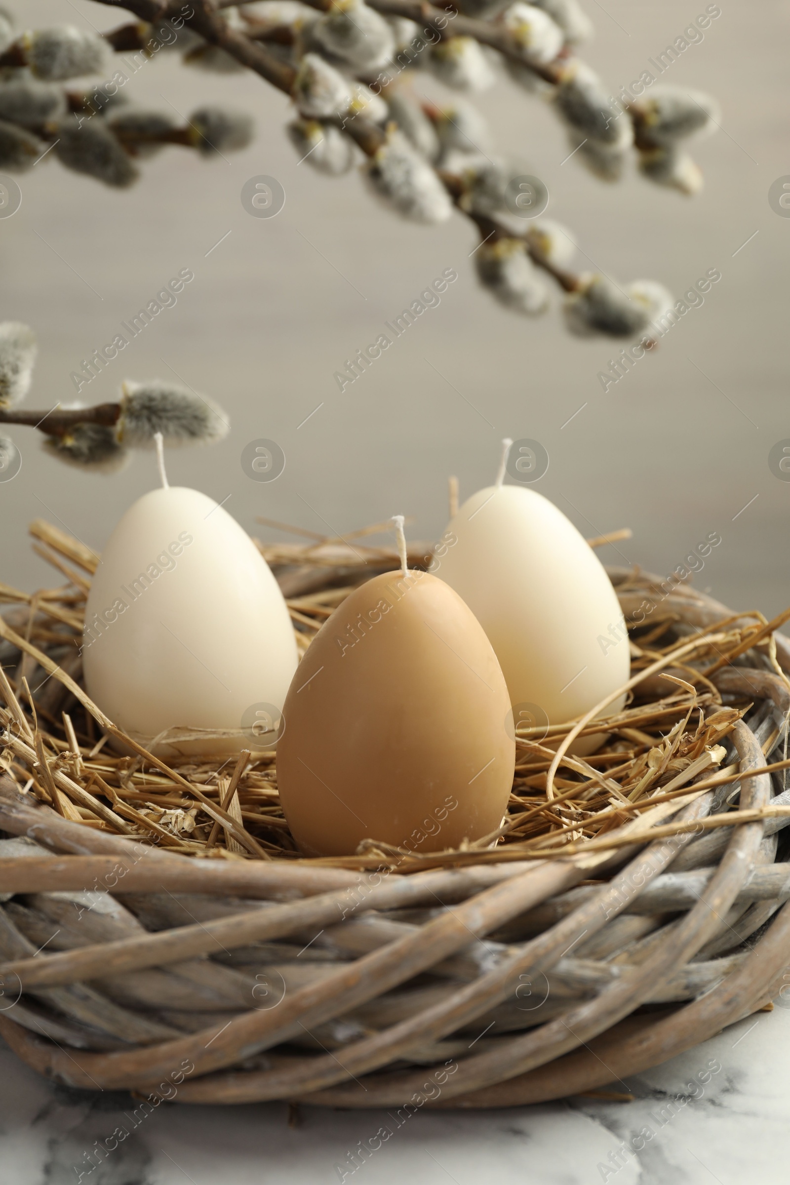 Photo of Egg-shaped candles in nest on white marble table and willow branches against light background, closeup. Easter decor