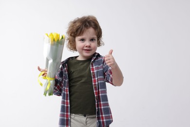 Photo of Cute little boy with bouquet of tulips showing thumbs up on white background