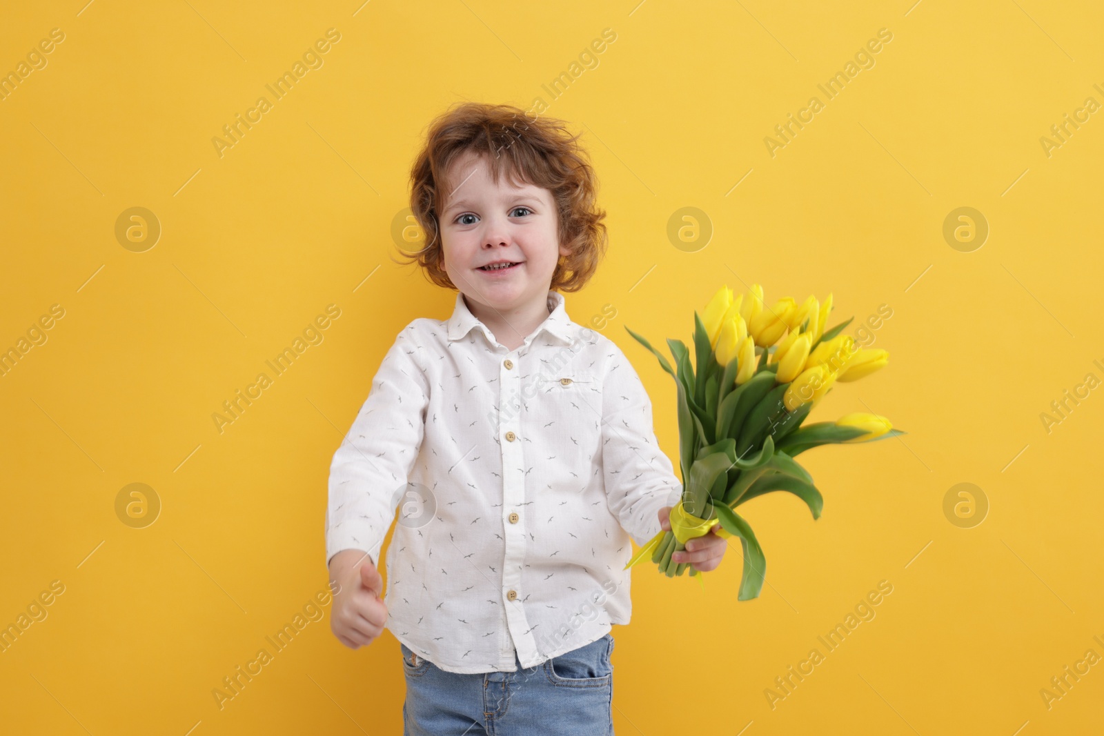 Photo of Cute little boy with bouquet of tulips showing thumbs up on yellow background