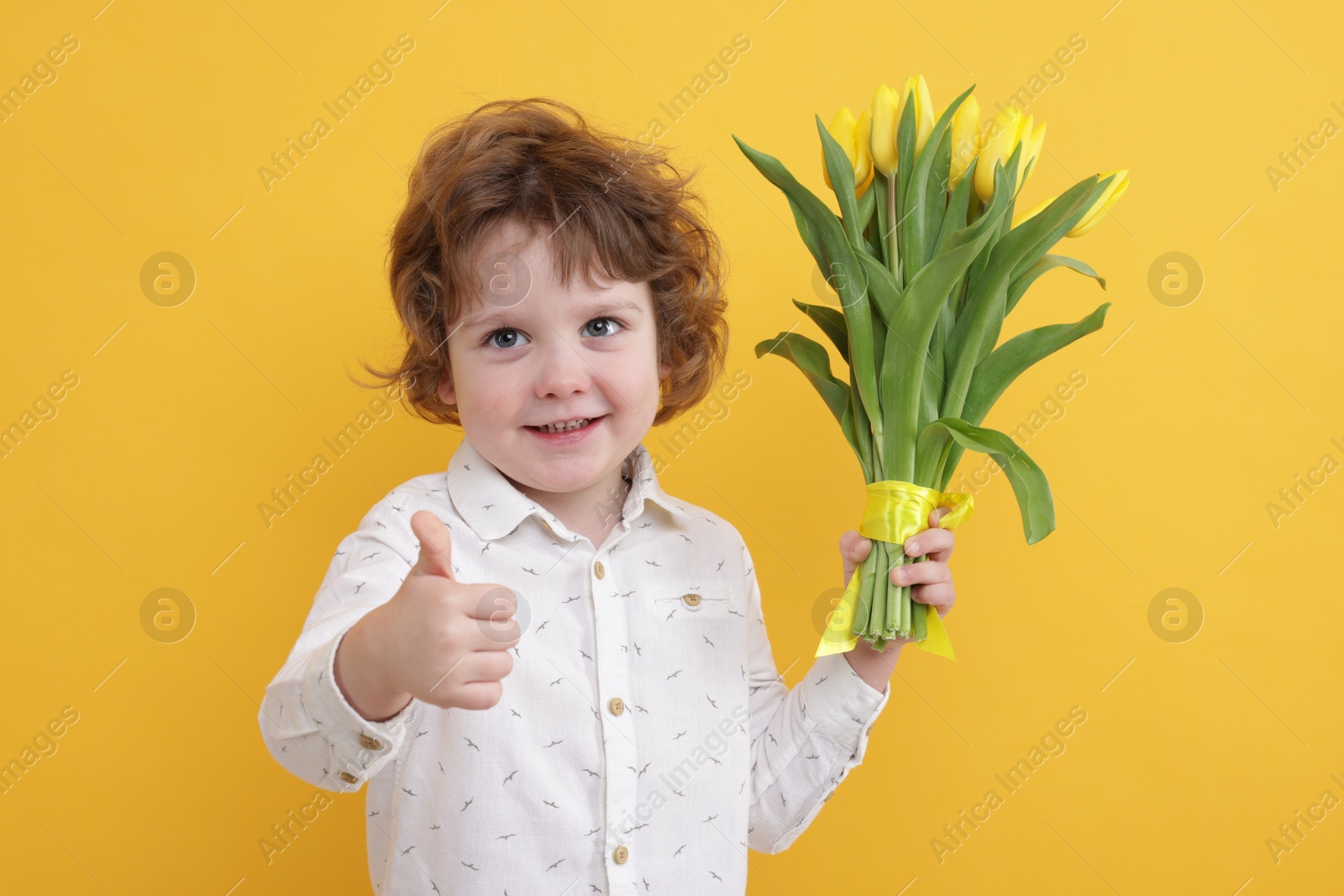 Photo of Cute little boy with bouquet of tulips showing thumbs up on yellow background