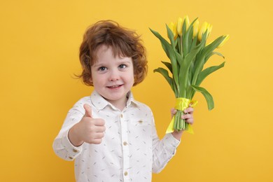 Photo of Cute little boy with bouquet of tulips showing thumbs up on yellow background