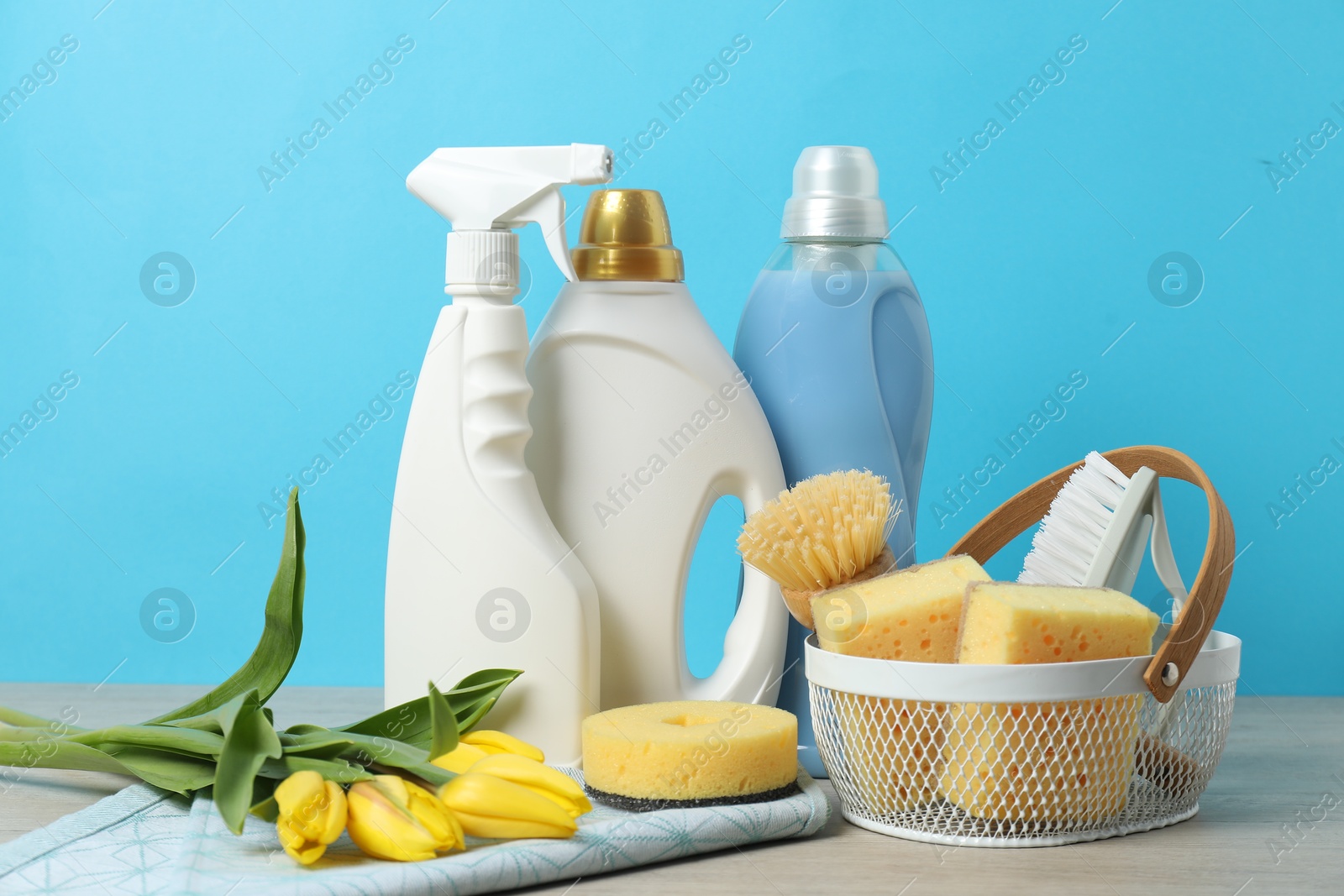 Photo of Spring cleaning. Detergents, supplies and tulips on white wooden table against light blue background