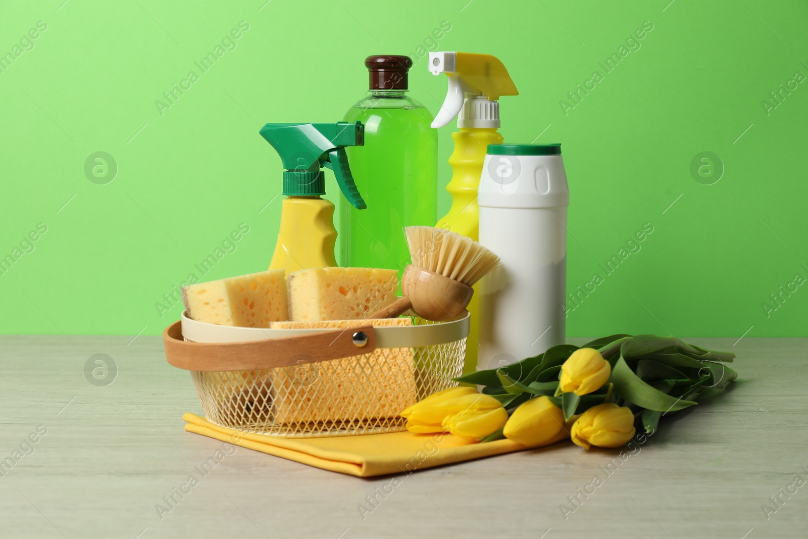 Photo of Spring cleaning. Detergents, supplies and tulips on white wooden table against green background