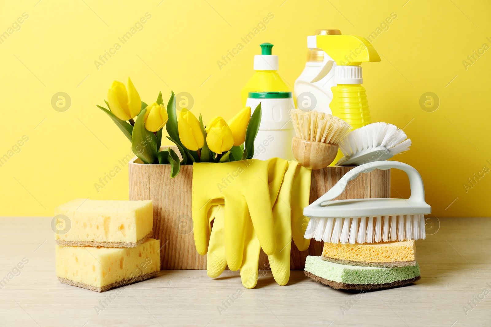 Photo of Spring cleaning. Detergents, supplies and tulips on white wooden table against yellow background