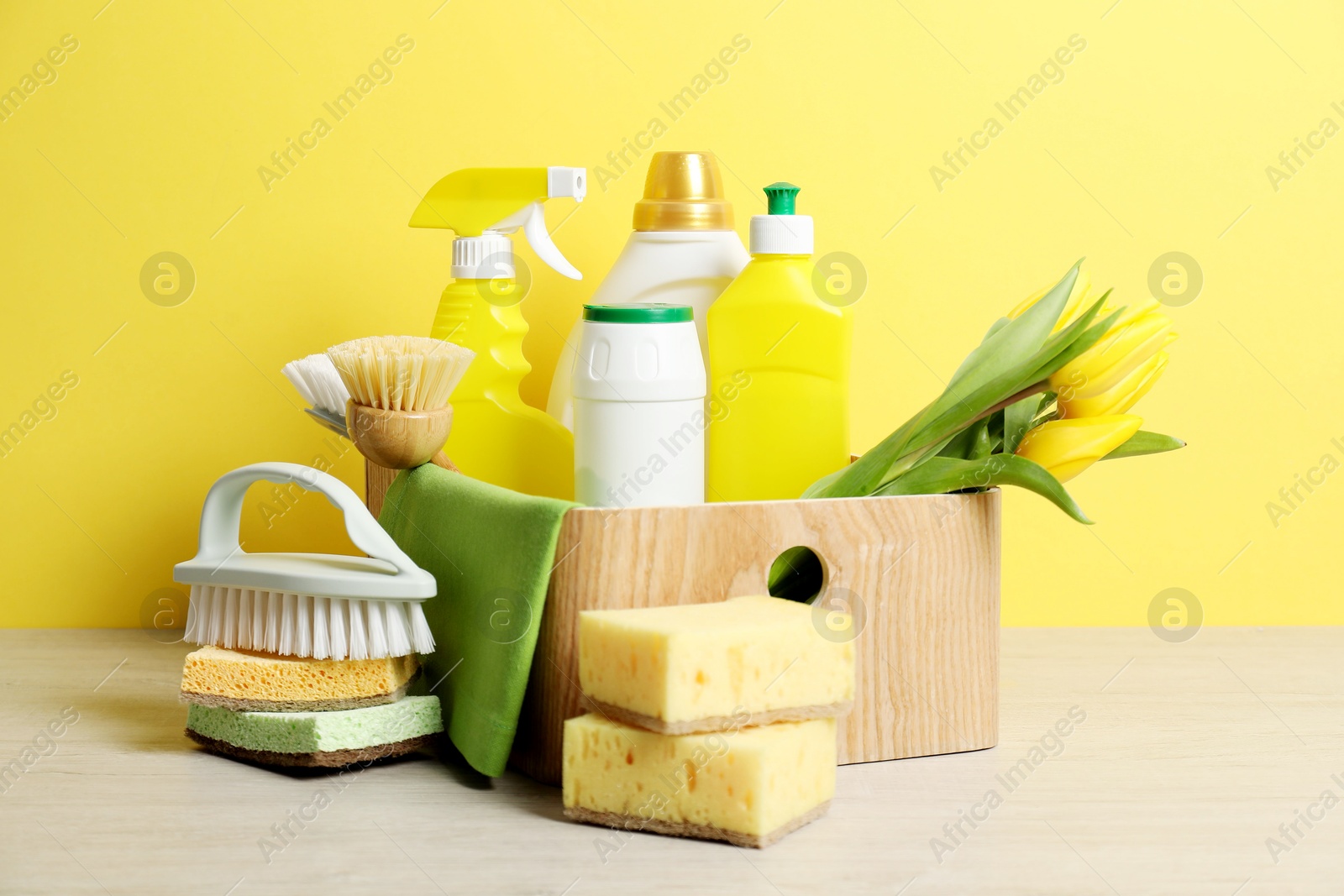 Photo of Spring cleaning. Detergents, supplies and tulips on white wooden table against yellow background