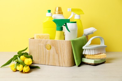 Photo of Spring cleaning. Detergents, supplies and tulips on white wooden table against yellow background