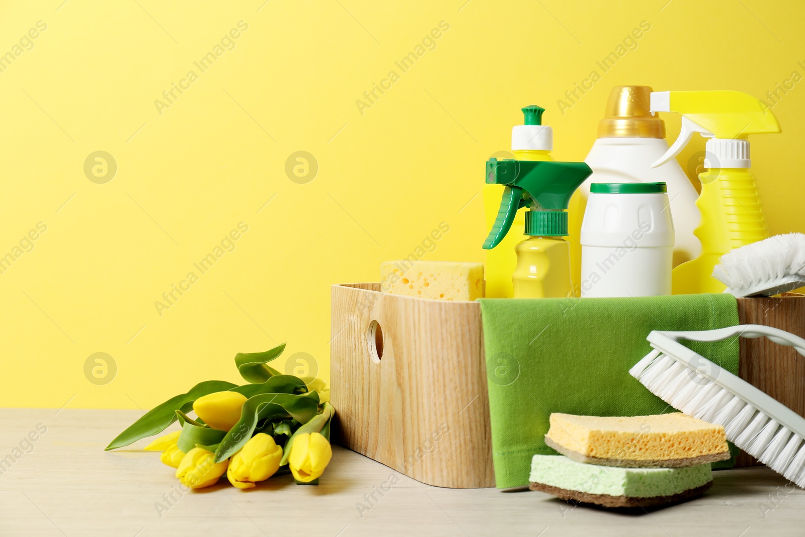 Photo of Spring cleaning. Detergents, supplies and tulips on white wooden table against yellow background, space for text