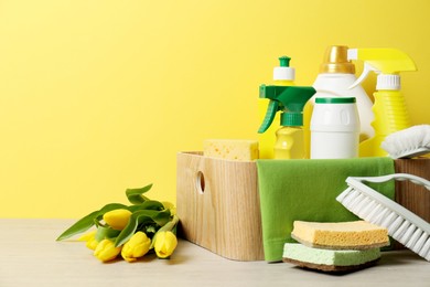 Photo of Spring cleaning. Detergents, supplies and tulips on white wooden table against yellow background, space for text