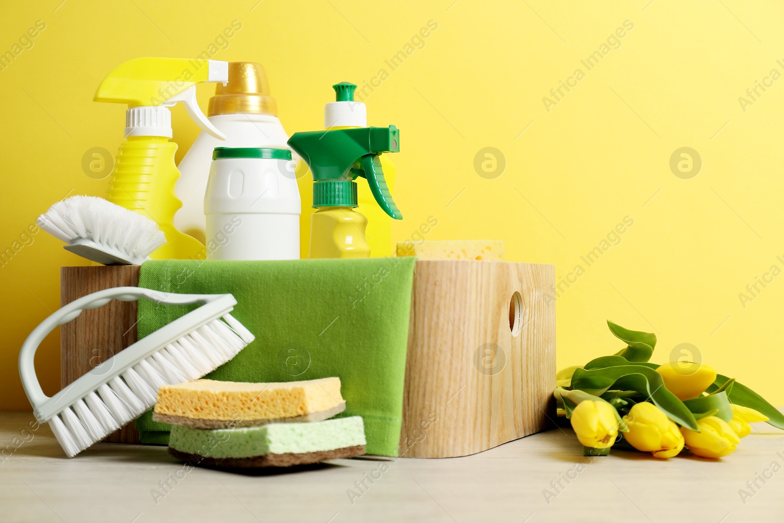 Photo of Spring cleaning. Detergents, supplies and tulips on white wooden table against yellow background