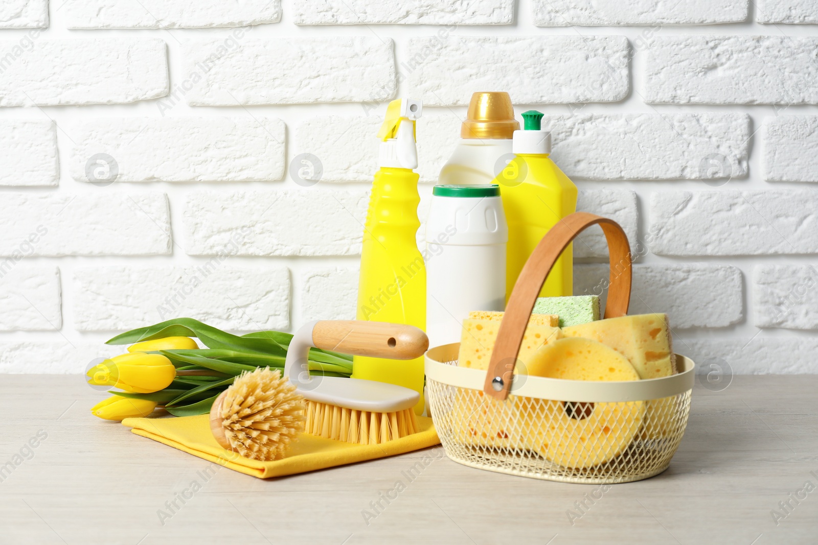 Photo of Spring cleaning. Detergents, supplies and tulips on white wooden table against brick wall