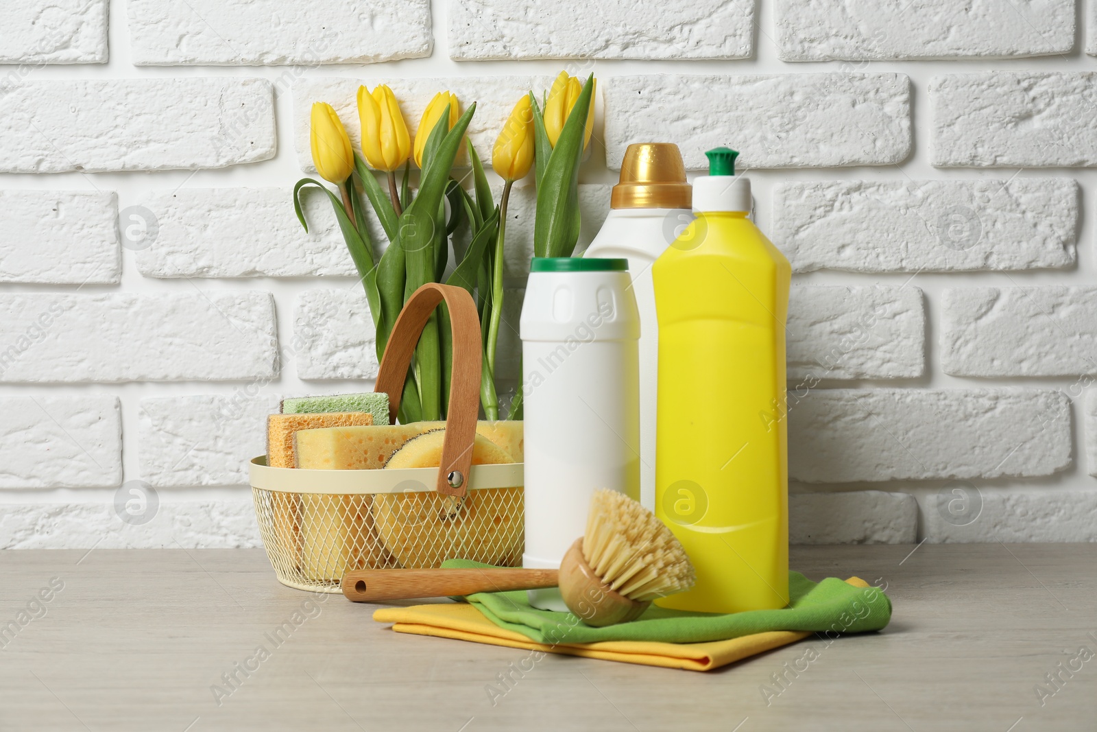 Photo of Spring cleaning. Detergents, supplies and tulips on white wooden table against brick wall