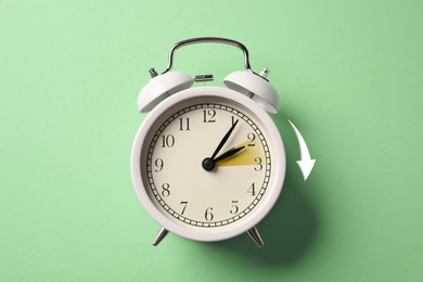 Image of Daylight saving time. White alarm clock on light green background, top view. Highlighted area and arrow showing time change by one hour forward