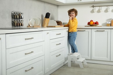 Photo of Little boy showing shush gesture while standing on step stool near countertop in kitchen