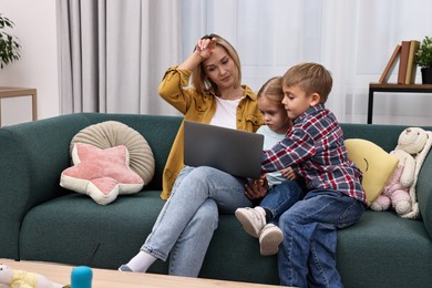 Photo of Tired housewife spending time with her kids and using laptop on couch at home